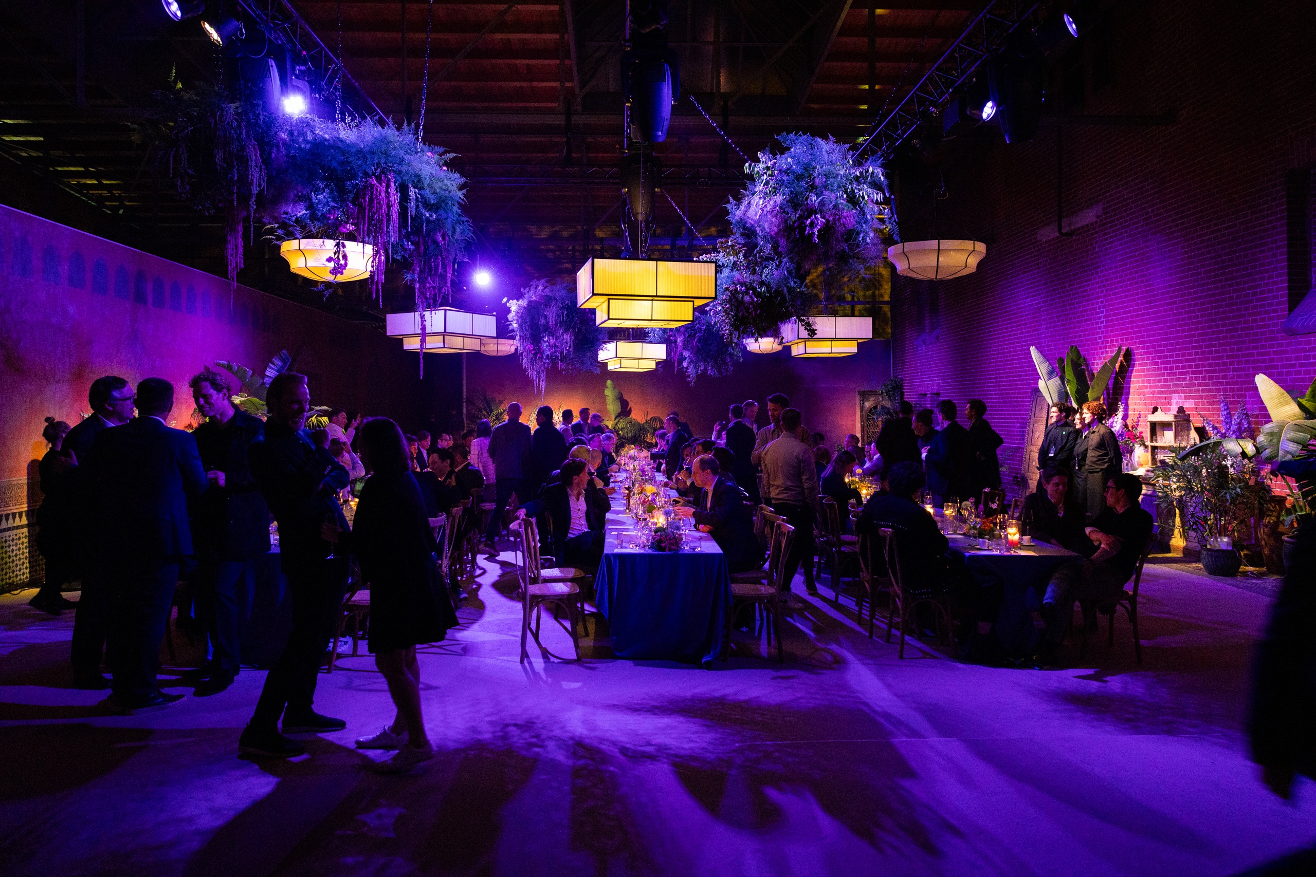 Guests at a long candlelit dinner under purple light in an industrial hall
